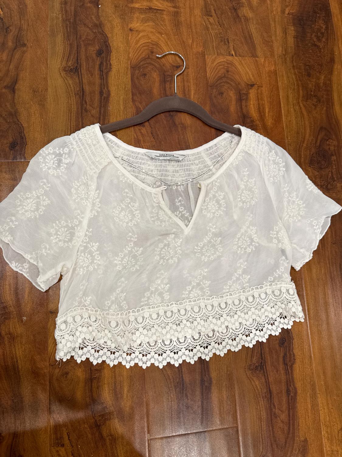 White lace blouse with short sleeves and floral details, hanging on a hanger over a brown wooden floor.
