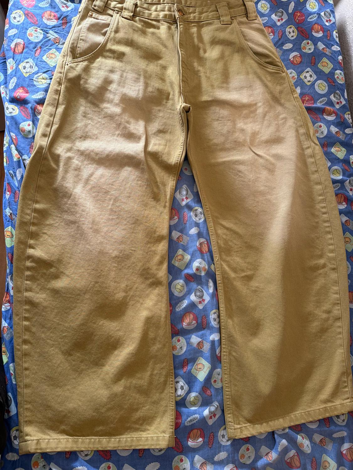 Mustard-coloured wide-leg trousers on a blue sheet with a football and ‘BOLOS’ text print. Relaxed style.