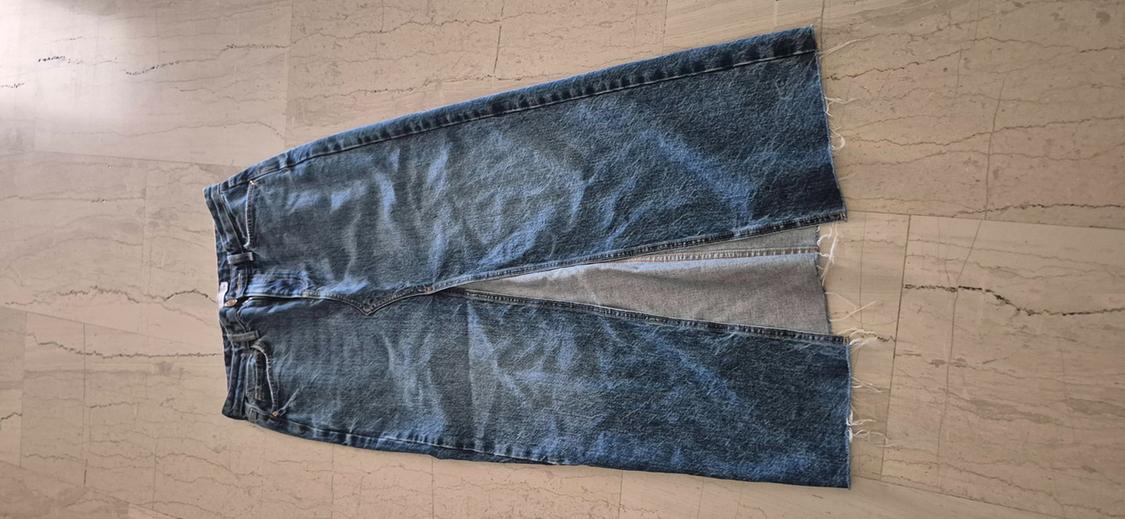 Long blue denim skirt with a front slit and frayed hem, laid out on a light-tiled floor.