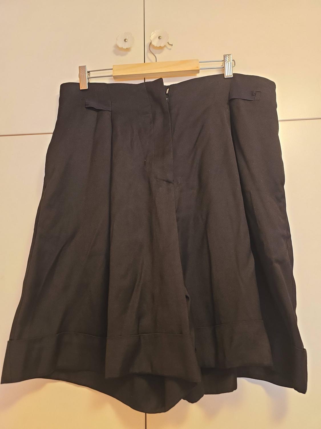 Wide black shorts with pleats and turn-up hems, hanging on a wooden hanger in front of a white wardrobe.