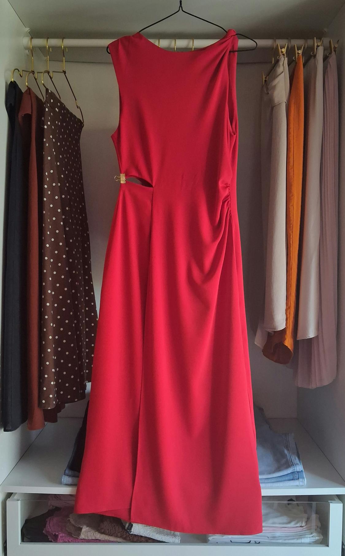Red sleeveless dress with side slit and gold clasp, hanging in a white wardrobe surrounded by clothes on hangers and folded garments.