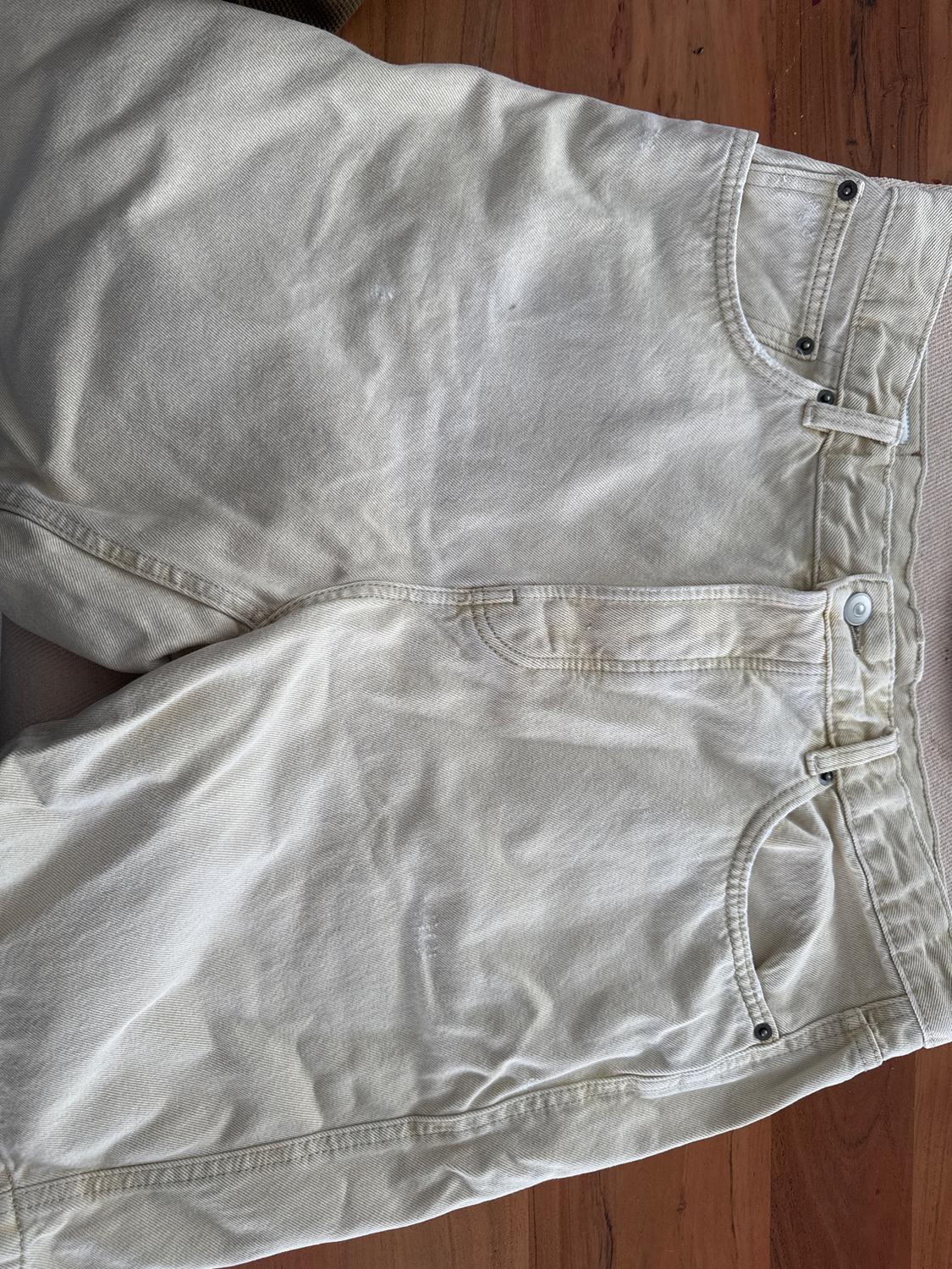 Creased beige denim shorts on a wooden floor. Visible front pockets and detailed stitching.