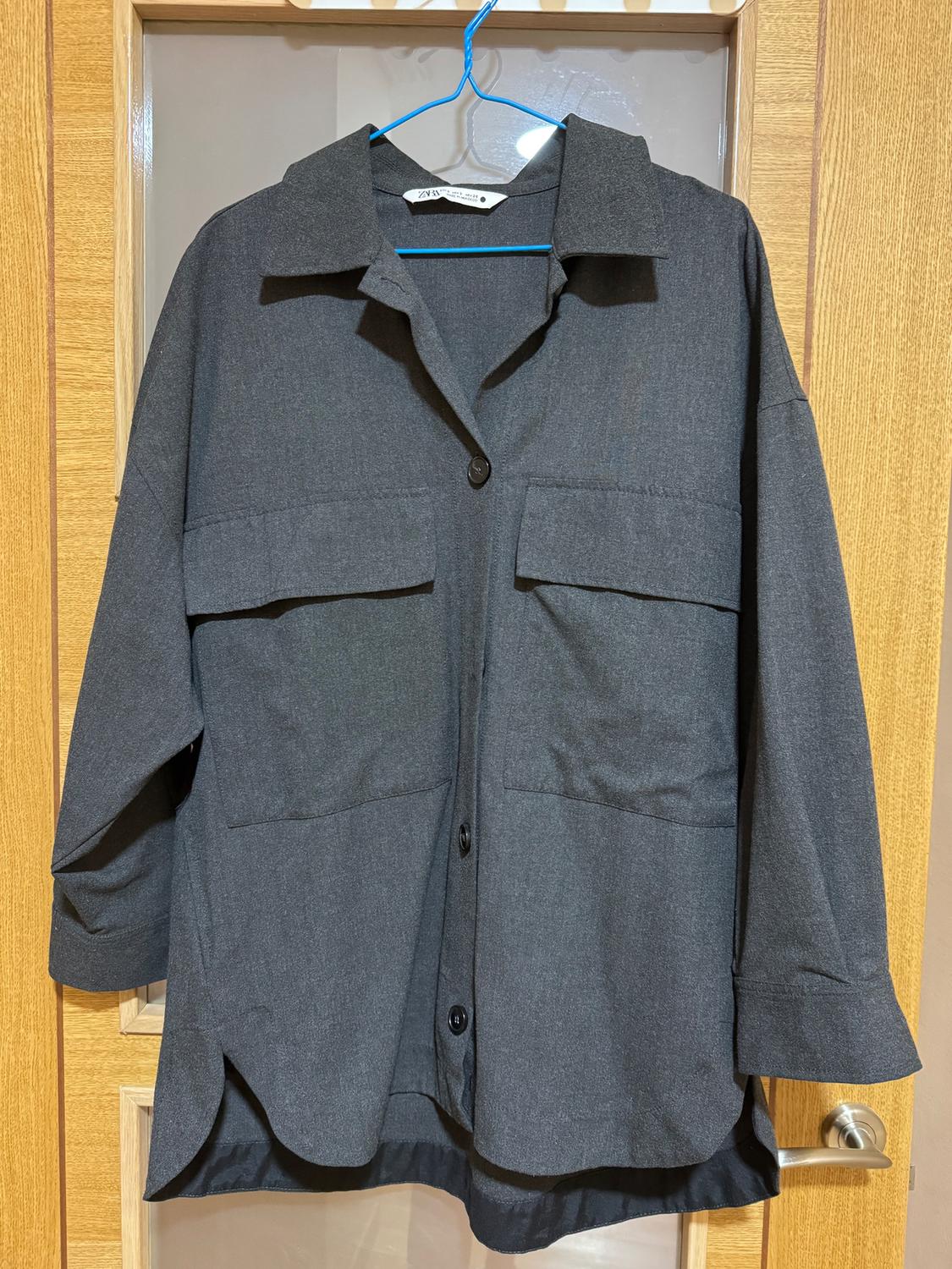 Grey, long-sleeved shirt hanging on a blue hanger over a wooden door. It has two front pockets and black buttons.