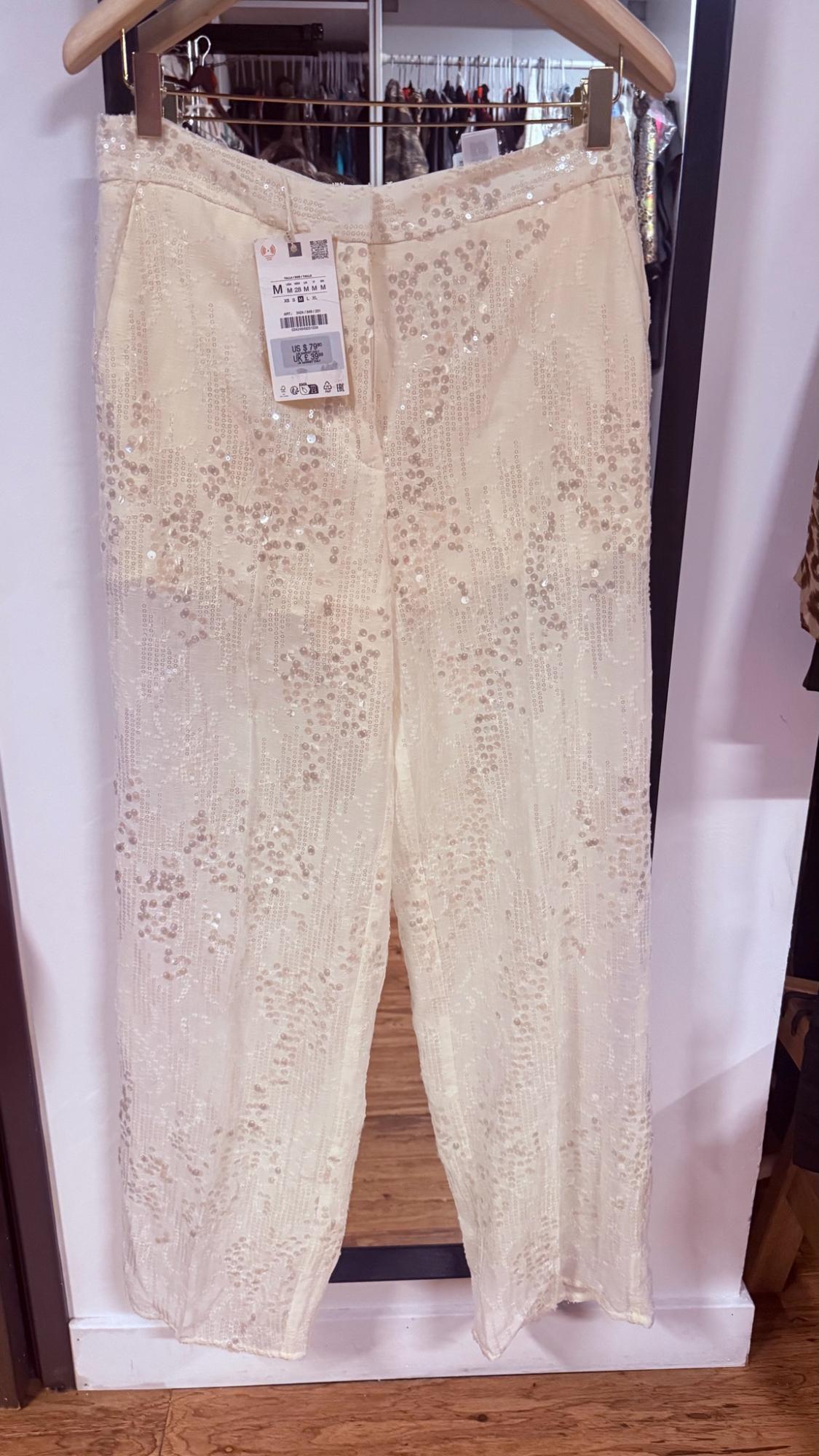 Cream-colored sequined pants hanging on a wooden hanger in front of a mirror.
In the reflection, you can see a coat rack.