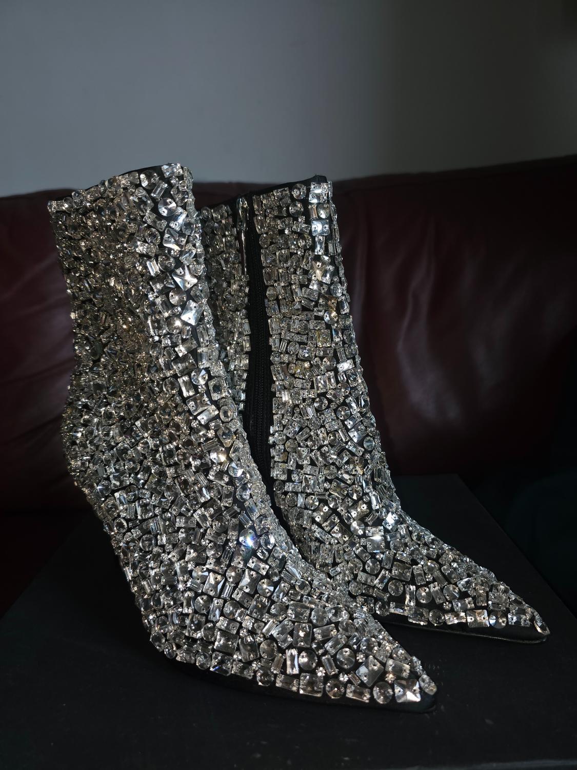 Pointed-toe heeled ankle boots covered in sparkling crystals, with a black zip, on a black box against a dark sofa background