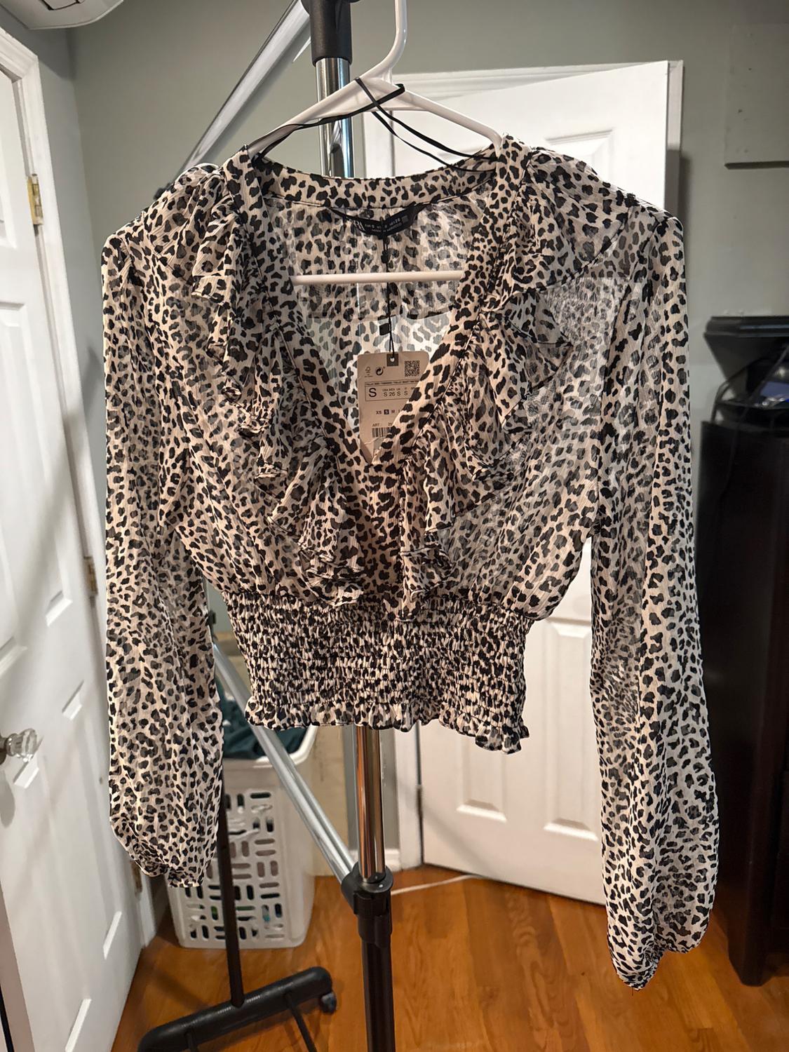 Long-sleeved blouse with leopard print on a hanger, room background with white doors and wooden floor. Visible label.