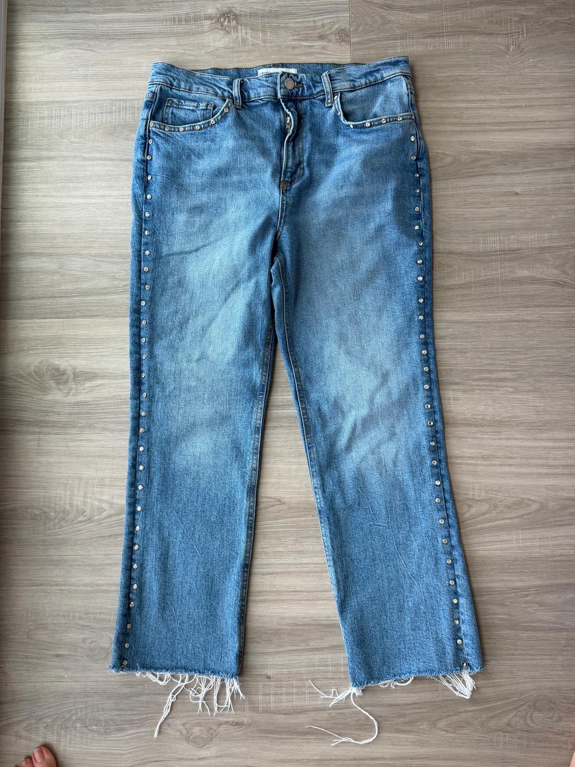 Blue jeans with studs on the sides and a frayed hem, placed on a light wooden floor.