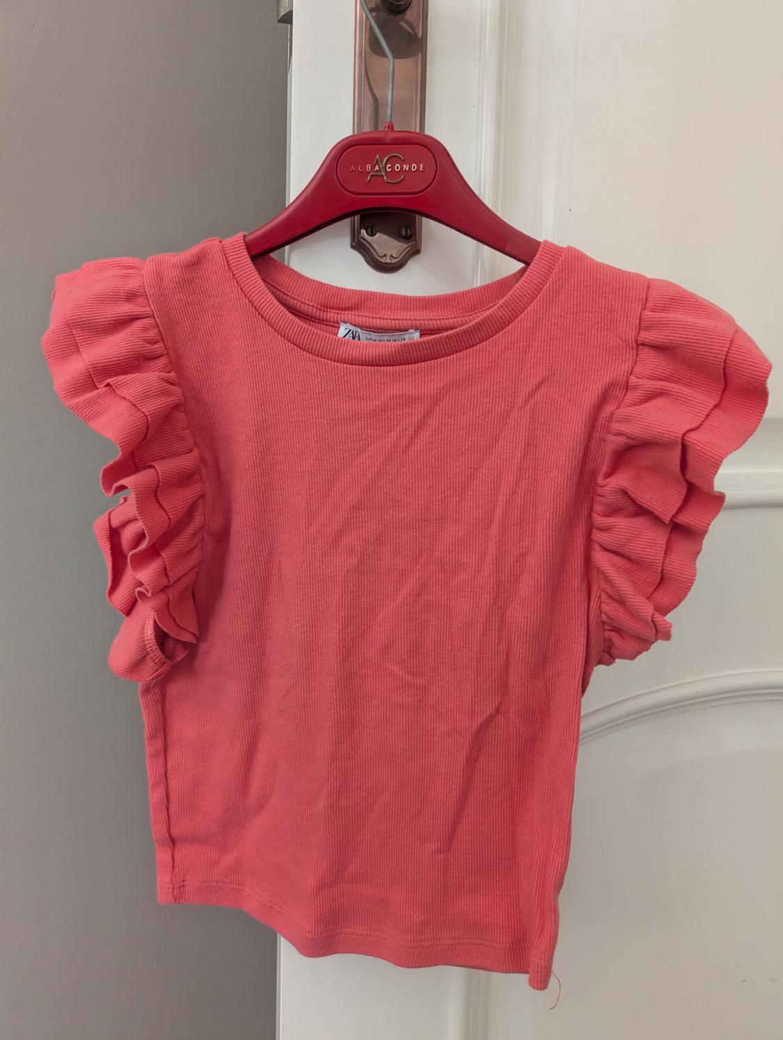 Pink T-shirt with ruffled sleeves, hanging on a red hanger with the "ALBA CONDE" logo on a white door.