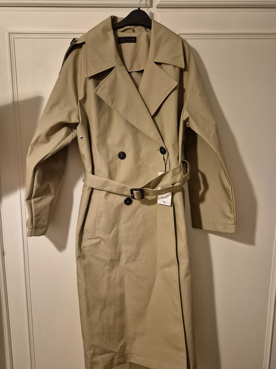 Beige trench coat hanging on a black hanger, with a black belt and buttons, against a background of white doors.