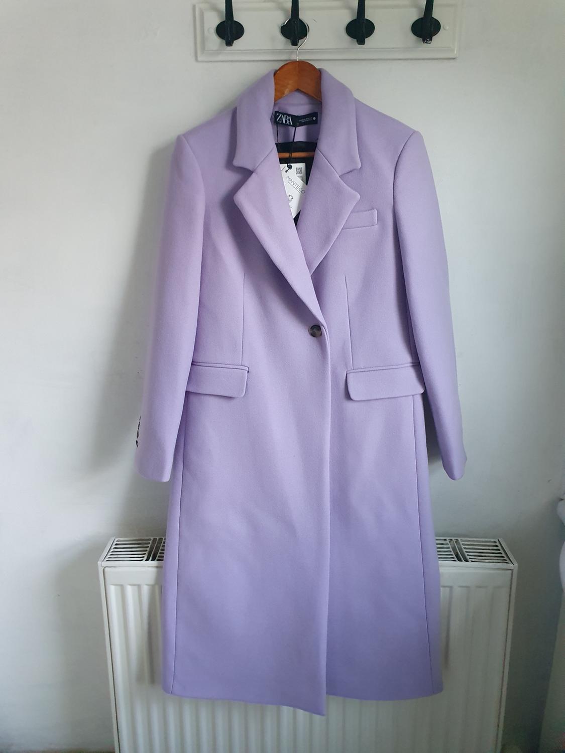 Long lilac coat hanging on a wooden hanger over a white radiator. Label visible inside the coat.