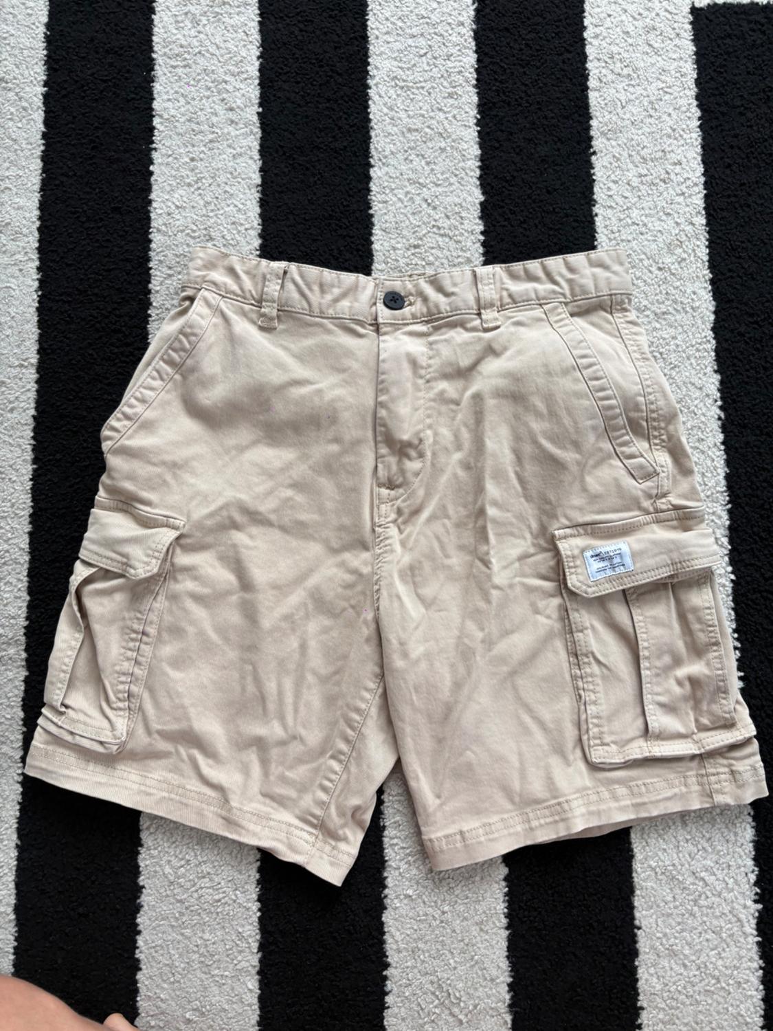 Beige shorts with cargo pockets on a black and white striped rug. Visible tag on the right pocket.