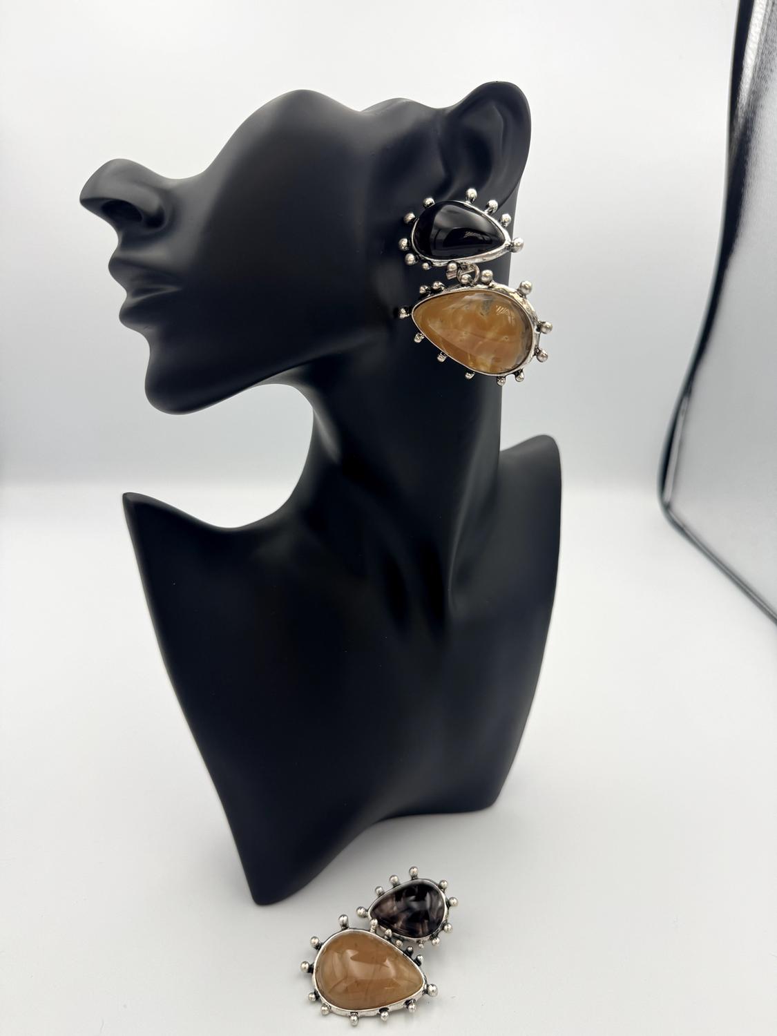 Black mannequin with large stone earrings in black and amber tones. White background with a silver bag to the right.