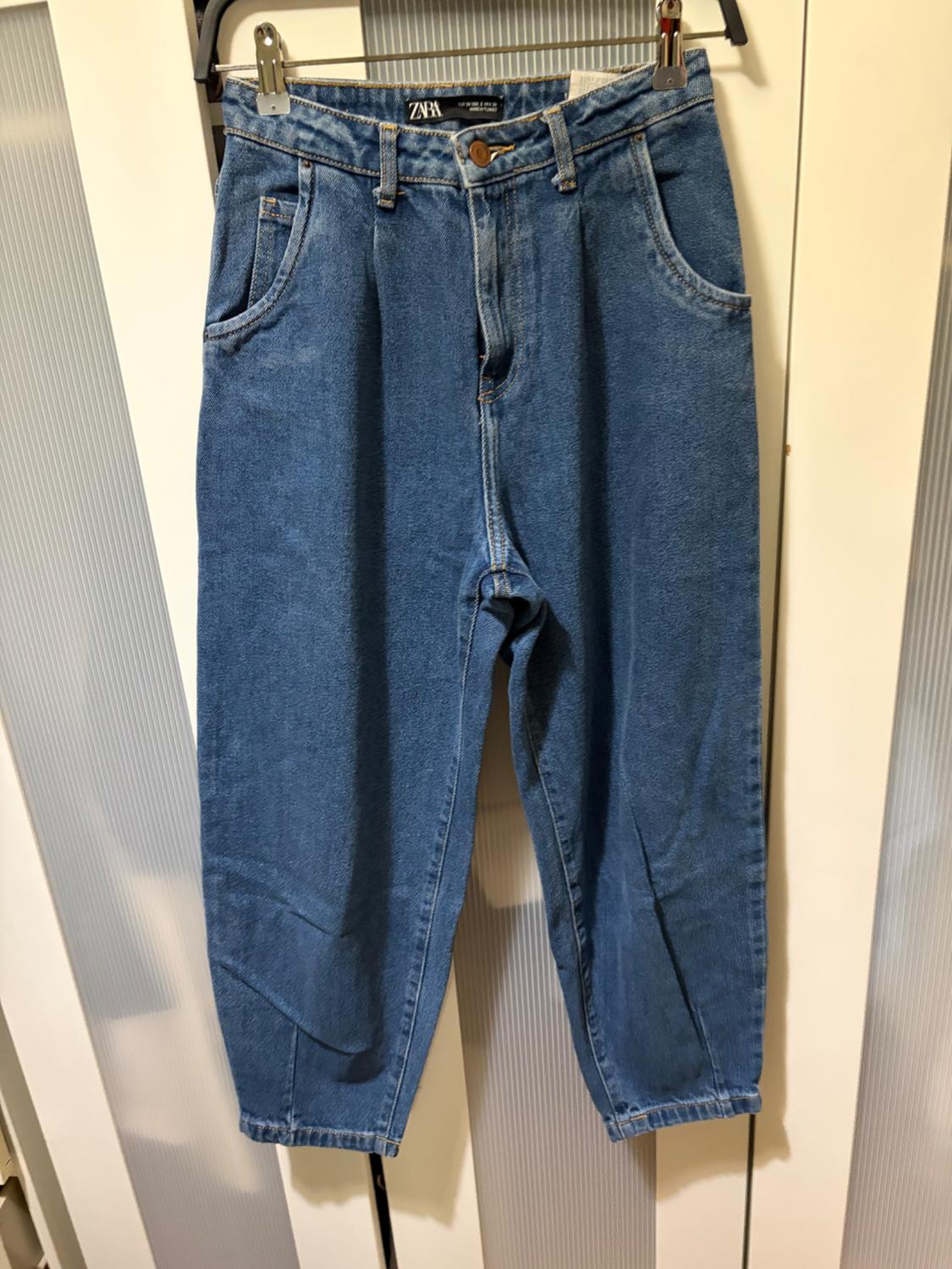 Blue denim jeans hanging on a hanger, with a visible label on the waistband that says "ZARA". Background of white doors and striped panels.