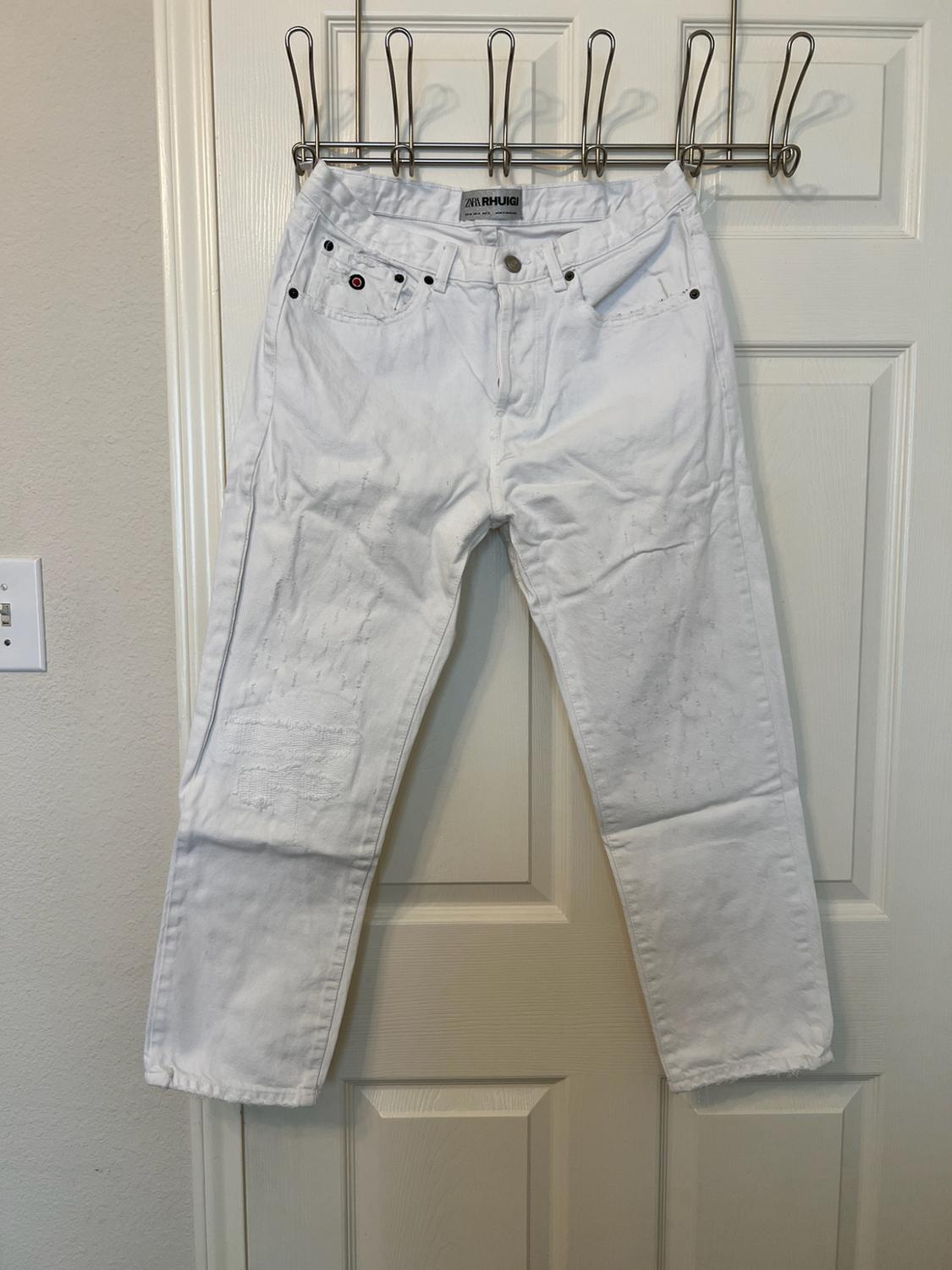 White denim pants with distressed details hanging on a door. Visible tag with the text ZARA RHUIGI.