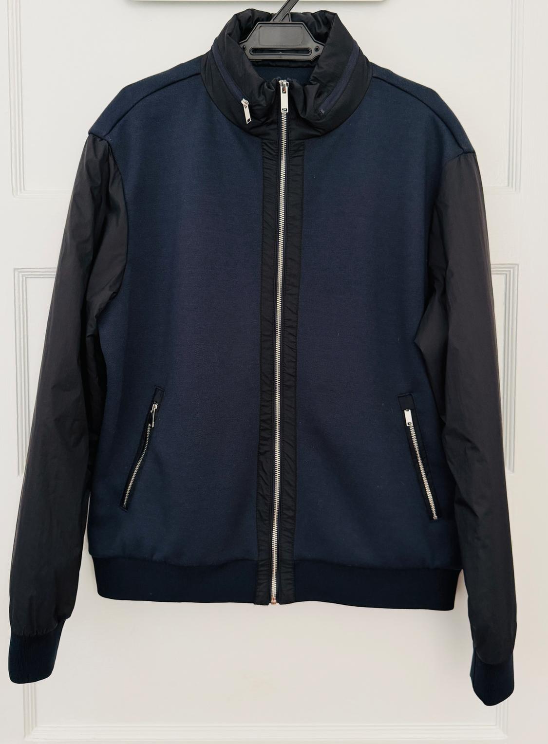 Navy blue jacket with black sleeves and silver zippers, hanging from a black hanger over a white paneled door.