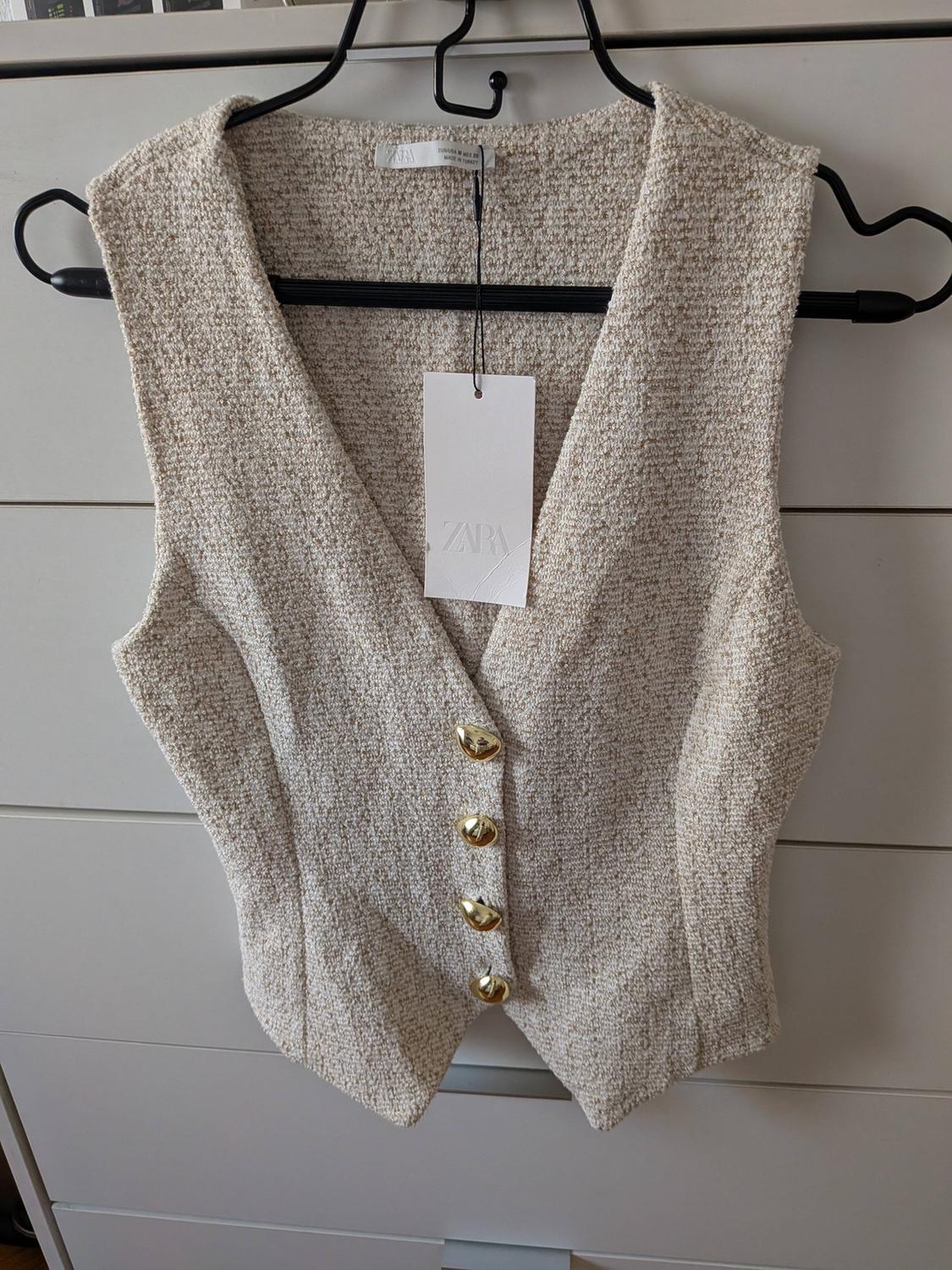 Textured beige V-neck waistcoat with gold buttons on a black hanger above white drawers. Label with the text ZARA.