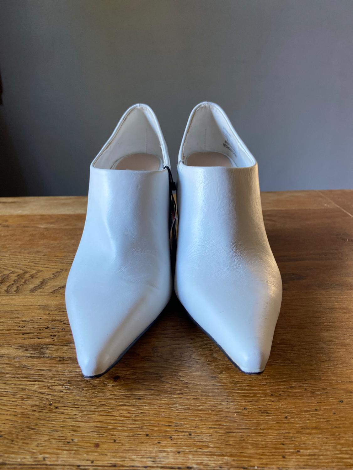 Pair of white pointed-toe ankle boots seen from the front, resting on a wooden surface with a neutral grey background.
