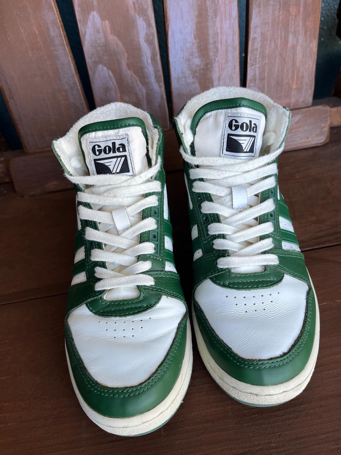 White and green athletic sneakers with laces, Gola brand visible on the tongue, on a brown wooden background.