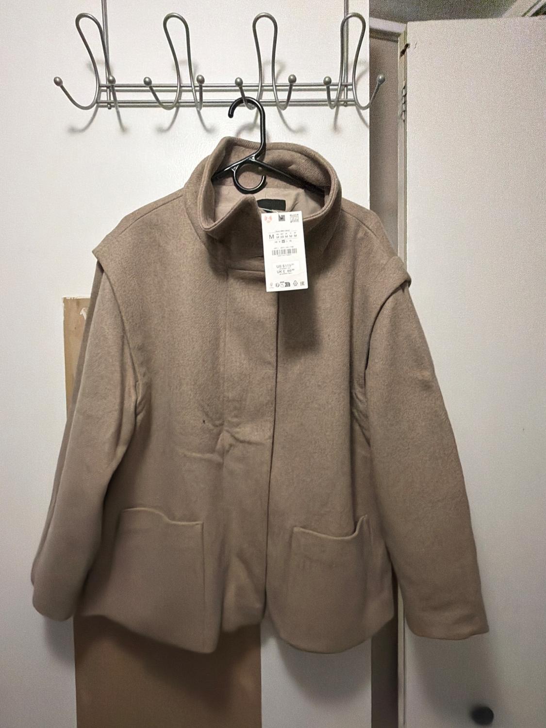 Beige coat with a high collar and front pockets, hanging on a coat hanger over a white door. It has a size M tag.