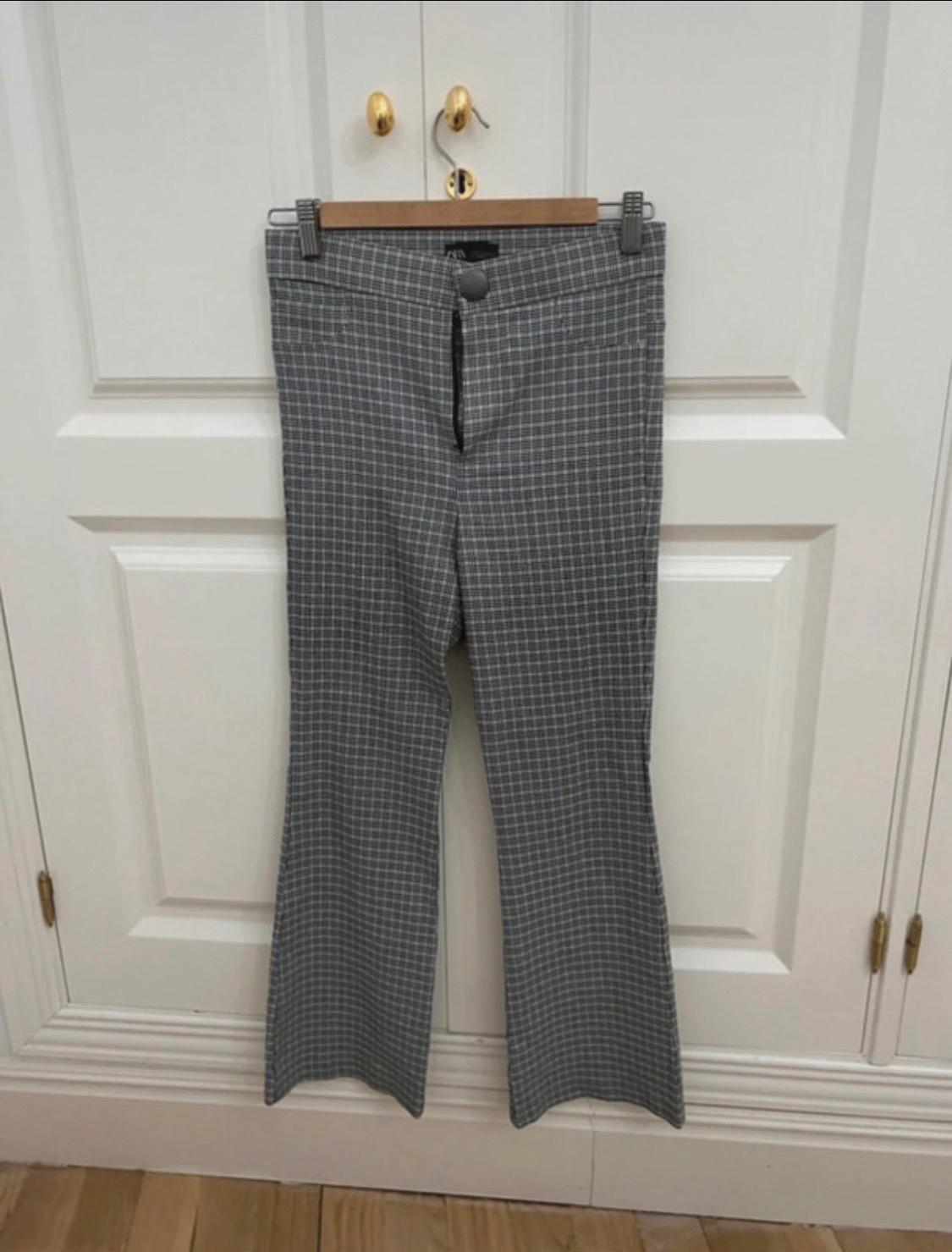 Grey and white checked trousers with a ZARA label, hanging from a wooden hanger on double white doors. Casual style.
