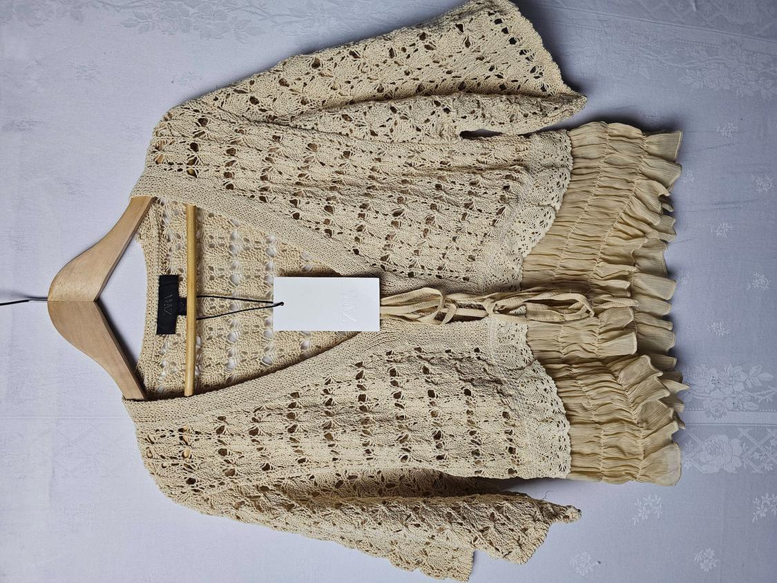 Beige crochet cardigan with fringe, hanging on a wooden hanger against a white background. White label visible.