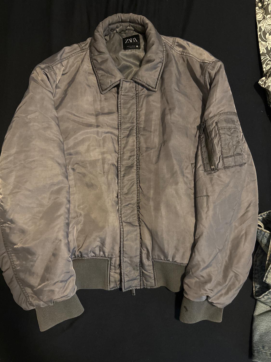 Grey padded jacket with zip and high neck on a black background. Zip pocket detail on the left sleeve.