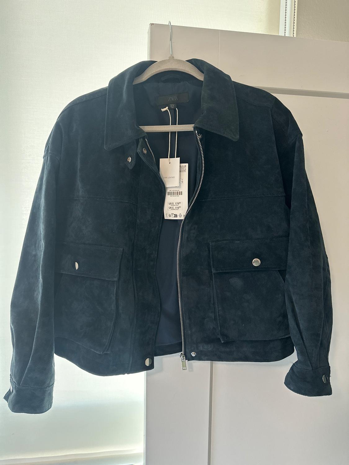 Black suede jacket hanging on a hanger, with a front zipper and buttoned pockets. Visible tag with price in dollars and pounds.