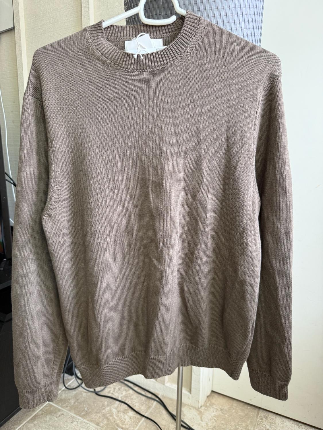 Light brown knitted jumper with a round neck and long sleeves, hanging on a white hanger in an indoor space with a tiled floor.
