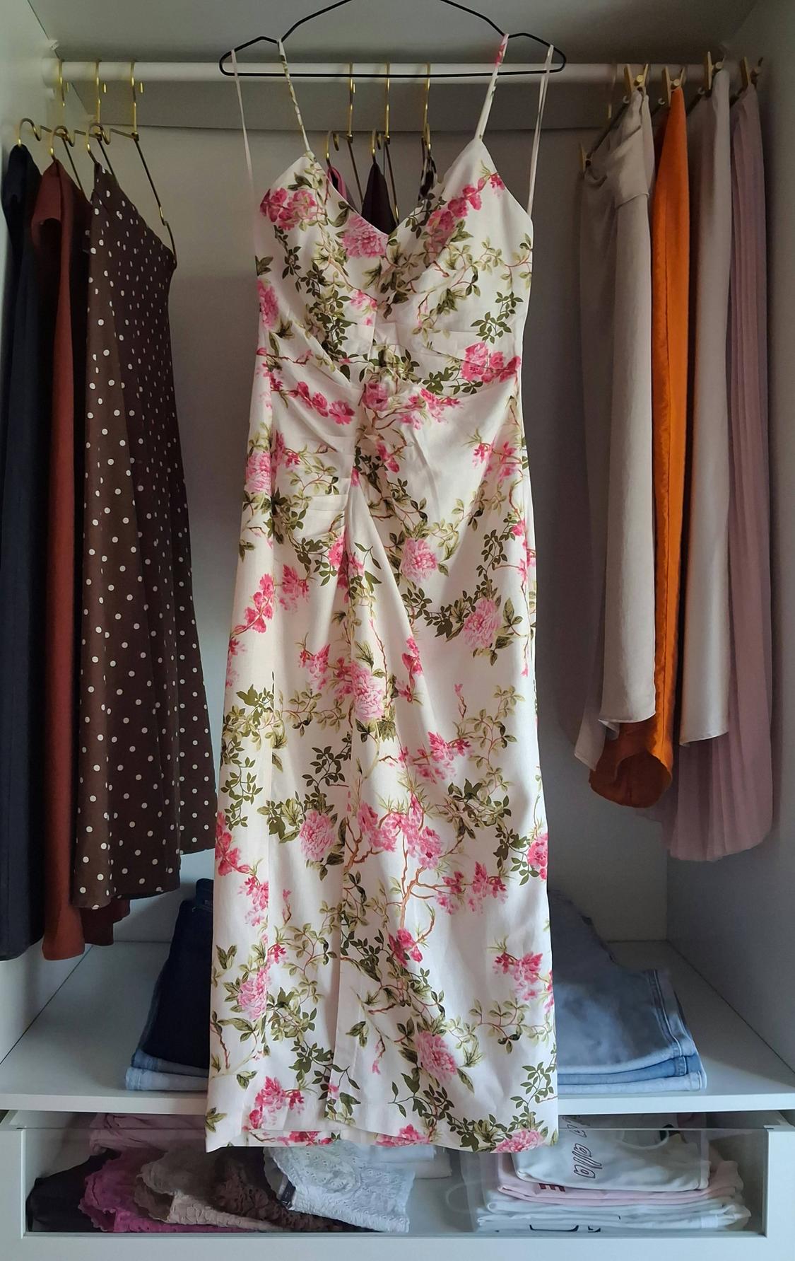 White dress with a pink floral print and straps, hanging in the centre of a wardrobe next to other garments and folded clothes on shelves.
