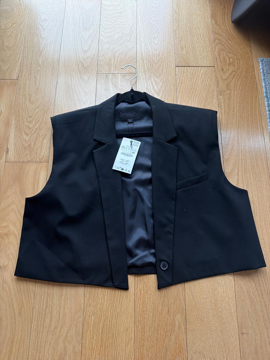 Sleeveless black vest hanging on a hanger over light wood flooring. Visible tag with product details.