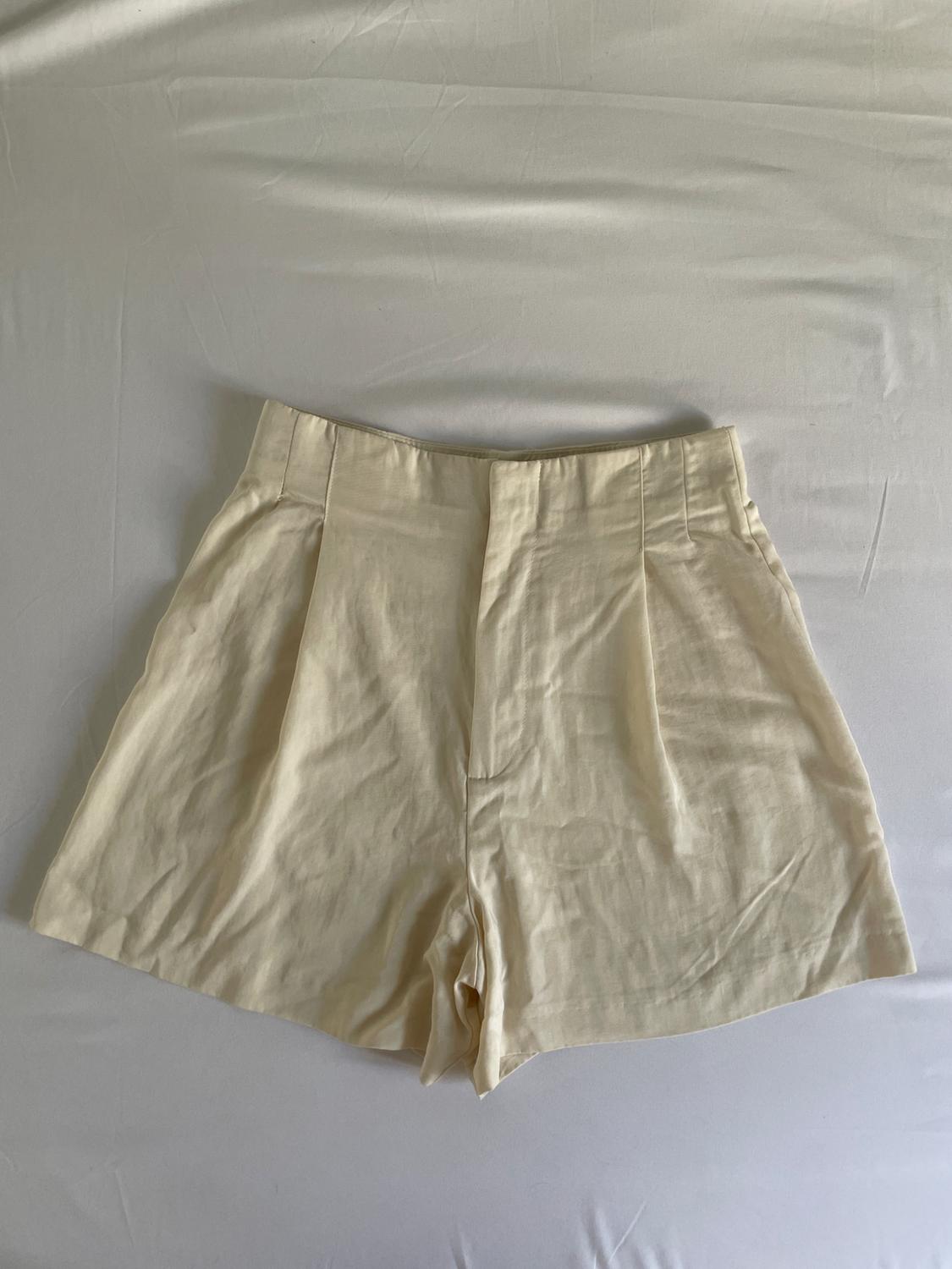 High-waisted beige tailored shorts with pleats, laid over a crumpled white fabric. Casual and minimalist style.