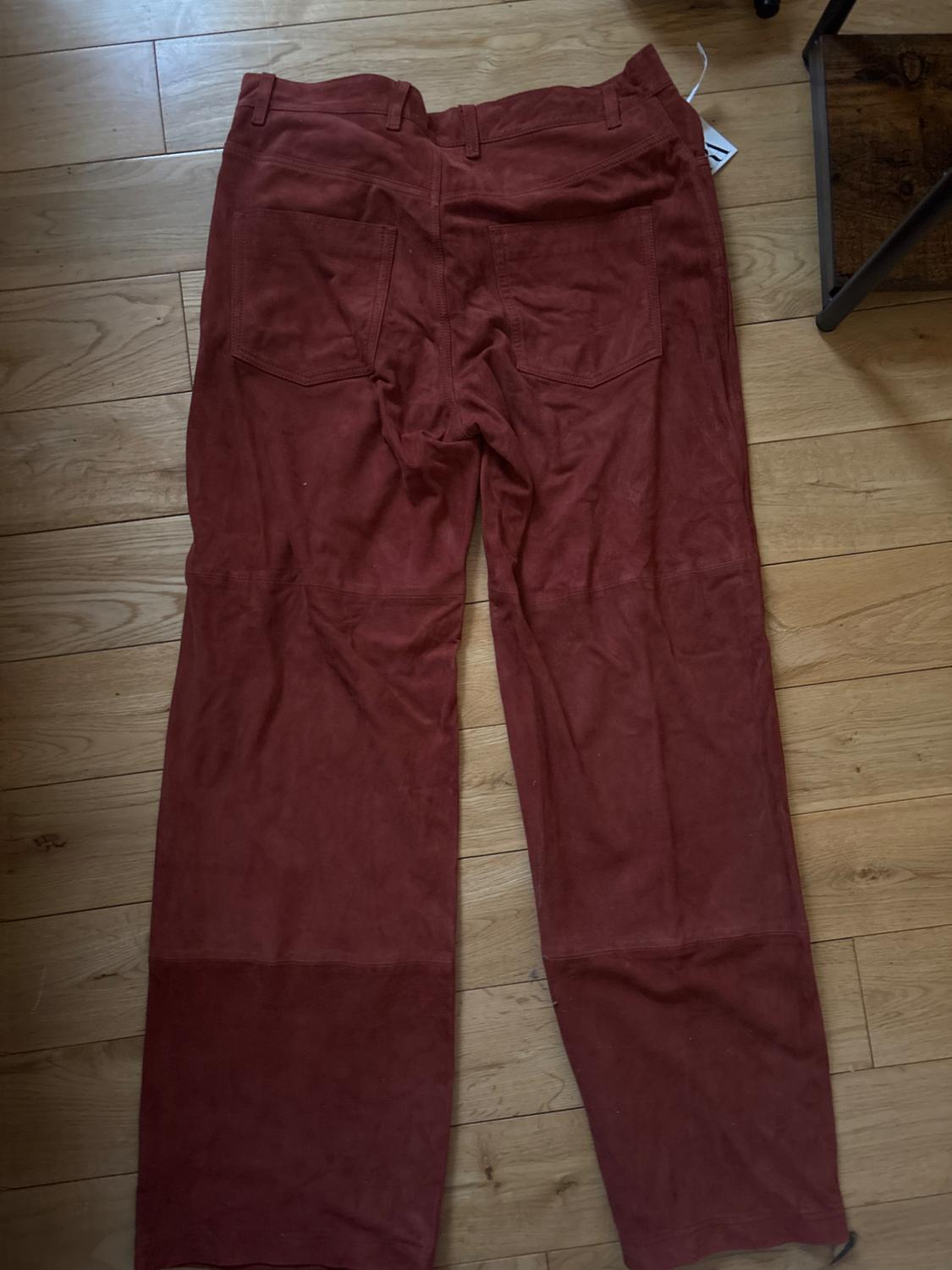 Red trousers made from thick fabric with back pockets, placed on a light wooden floor. Label visible at the top.