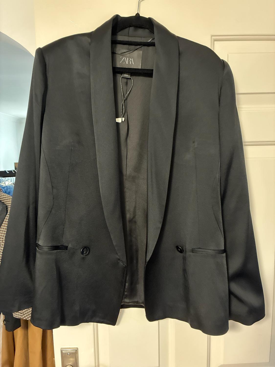 Satin-finish black blazer hanging on a hanger in front of a white door. ZARA label visible.