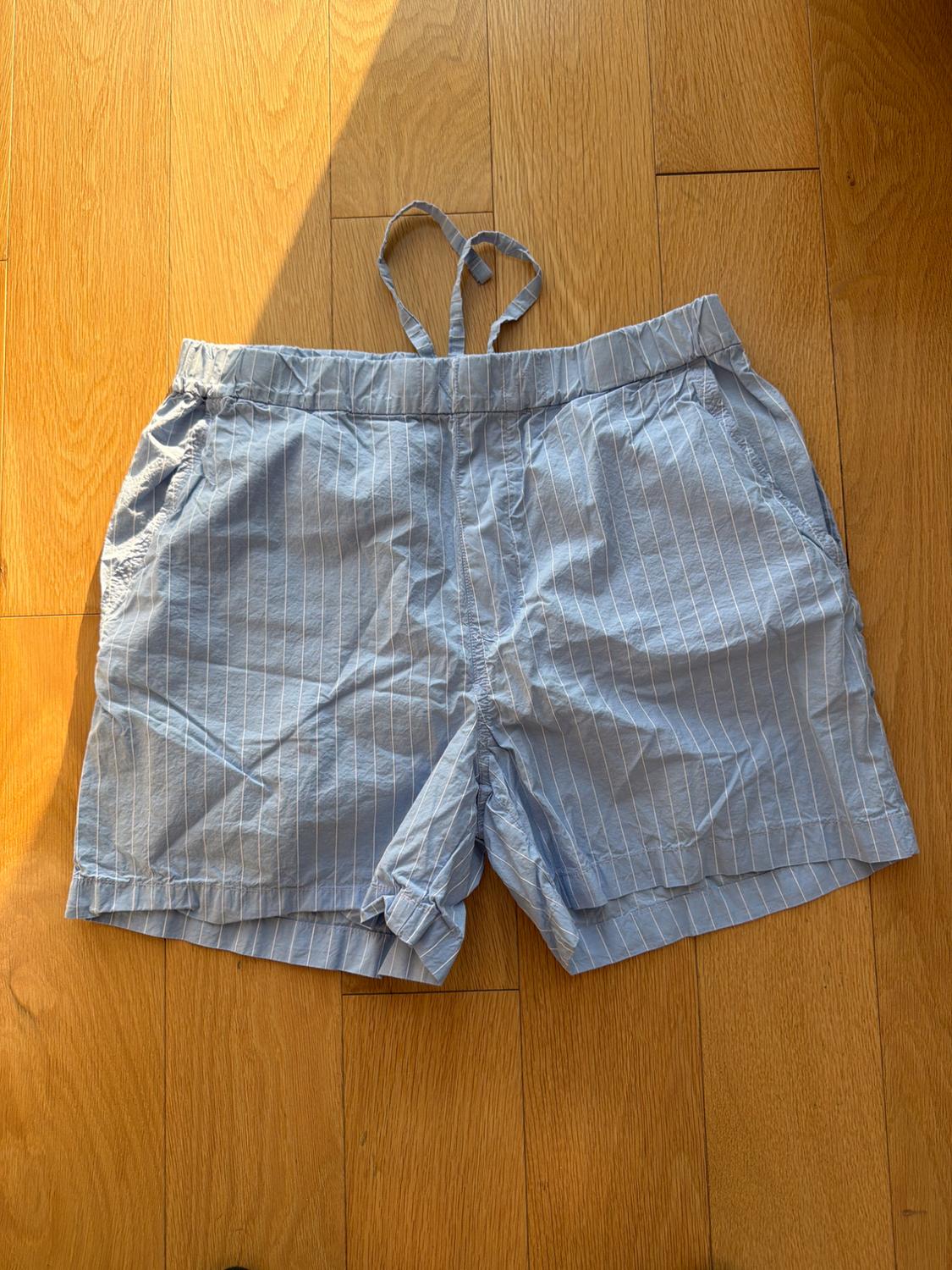 Blue shorts with white stripes on a light wooden floor, with an elastic waistband and adjustable drawstring. Natural light partially illuminates.