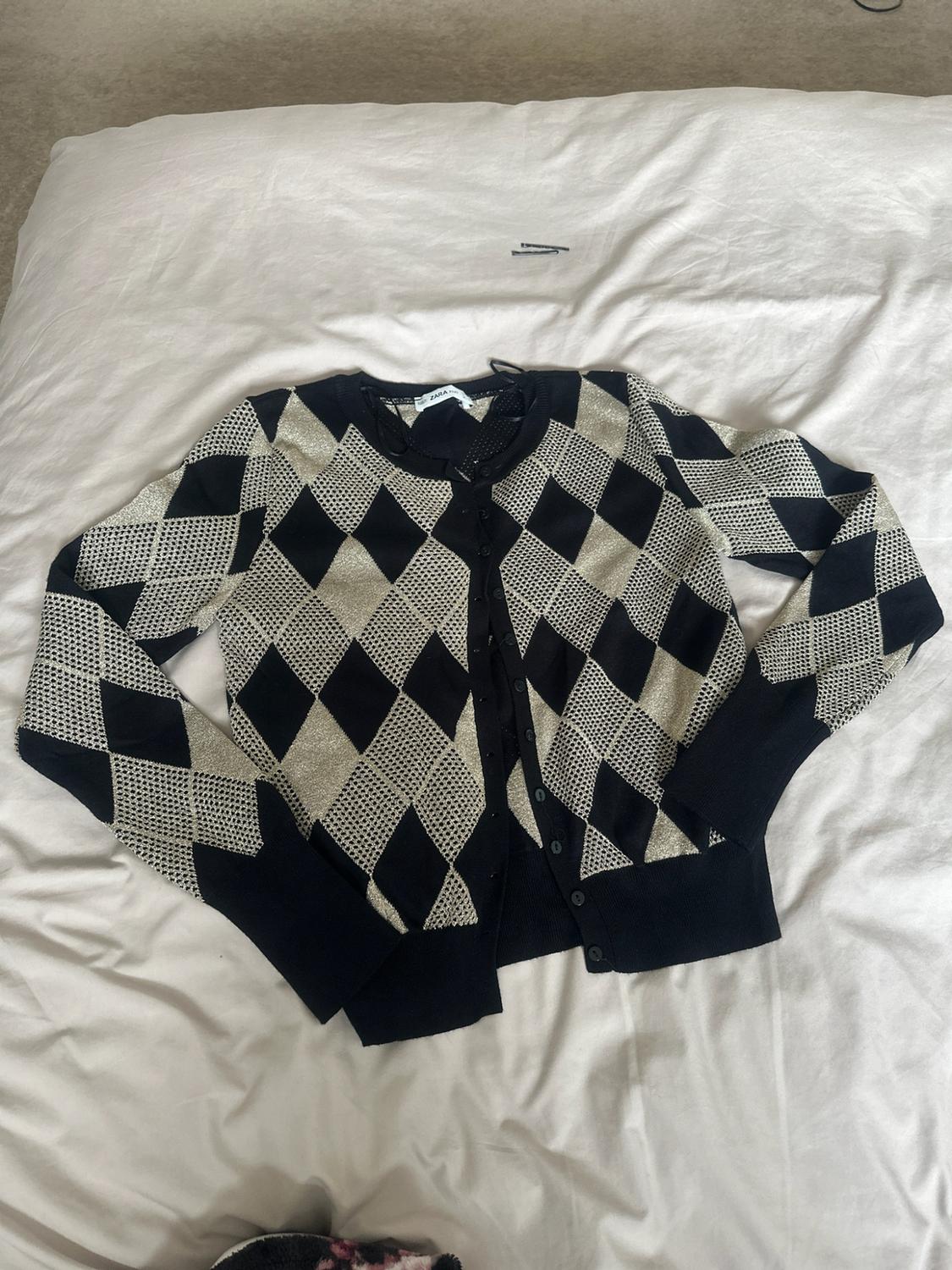 Black and beige argyle-pattern button-up cardigan, on crumpled white bedsheets. Relaxed atmosphere. Label text: ZARA.