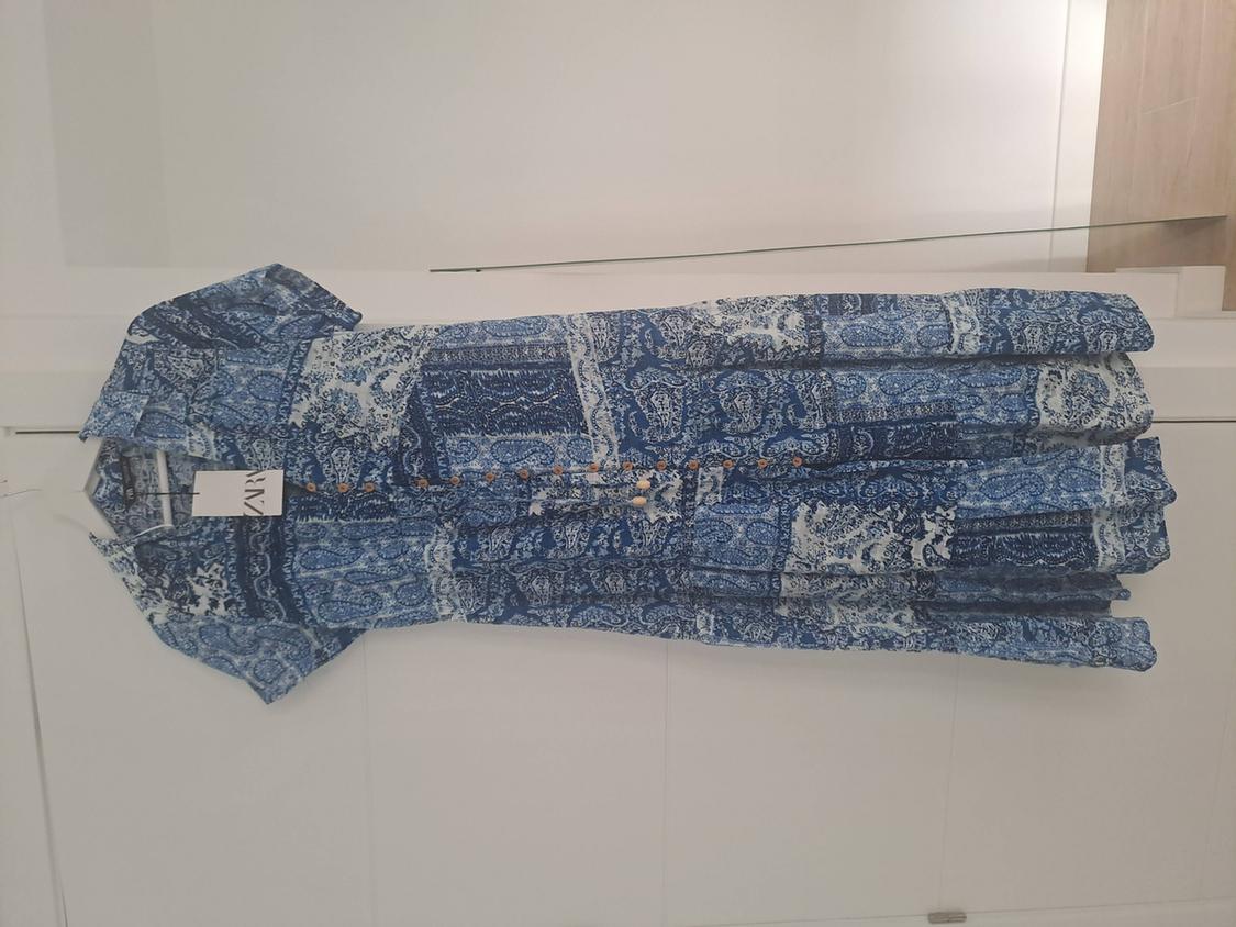 Long shirt dress with a blue and white print, short sleeves and buttons, hanging on a white door. Label with the text ZARA.