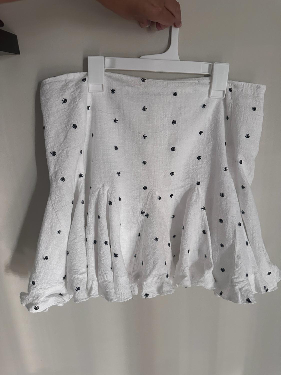 Short white skirt with small black flowers and a frilled trim, hanging from a hanger held by a hand in front of a light-coloured wall.