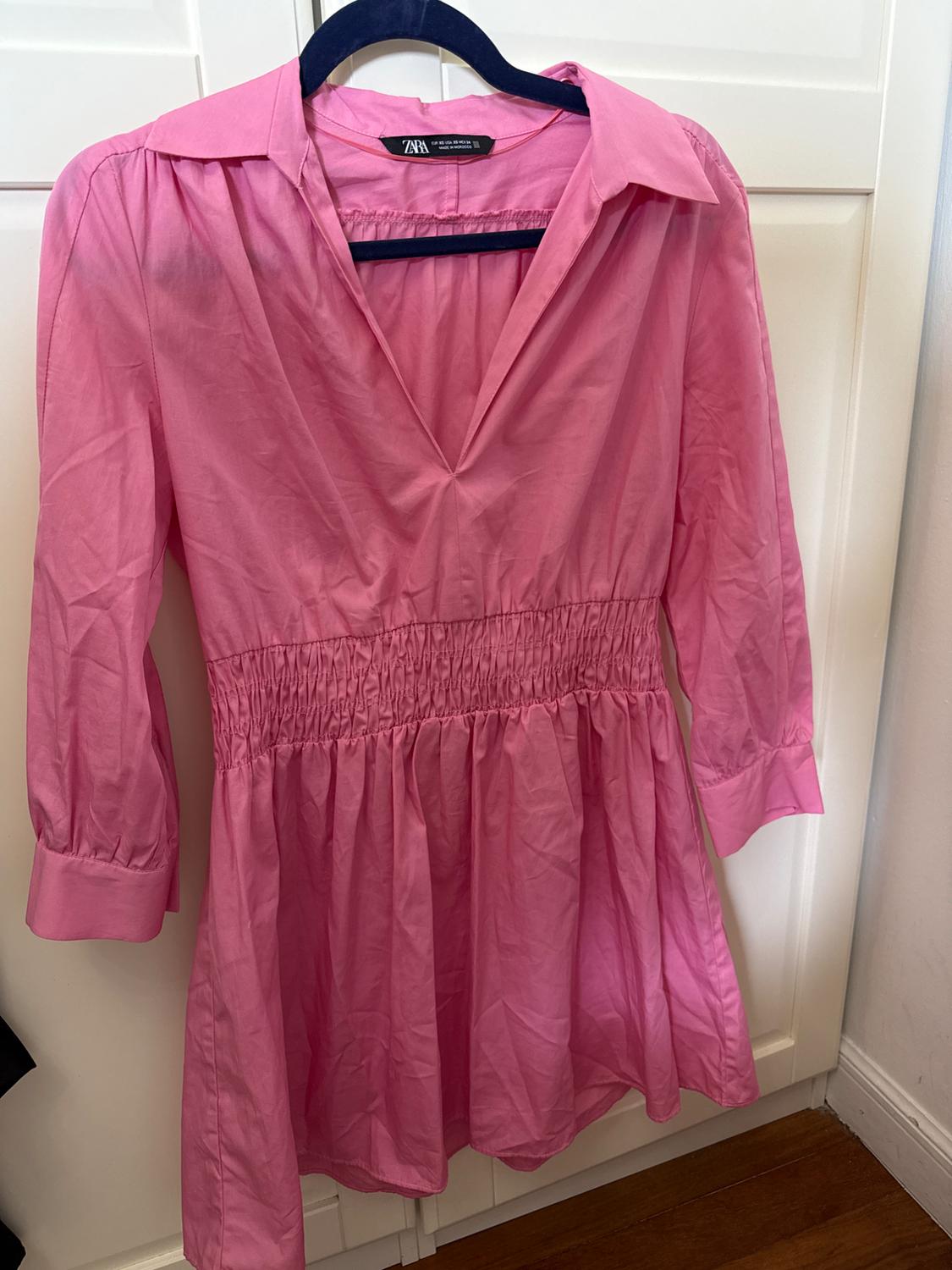 Pink long-sleeved dress with a lapel collar and elasticated shirred waist, hanging on a black hanger in front of white doors. ZARA text.