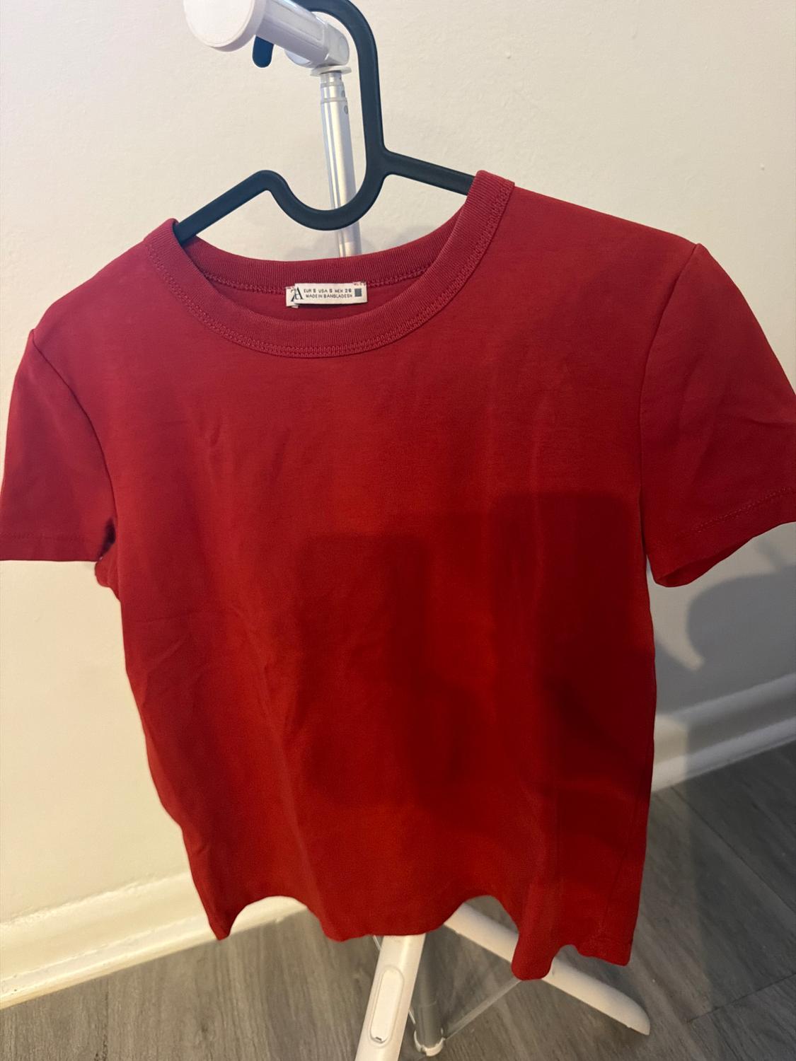 Red short-sleeved t-shirt hanging on a black hanger. White wall background and gray floor. Text: ZARA EUR S USA S MEX 26 MADE IN BANGLADESH.