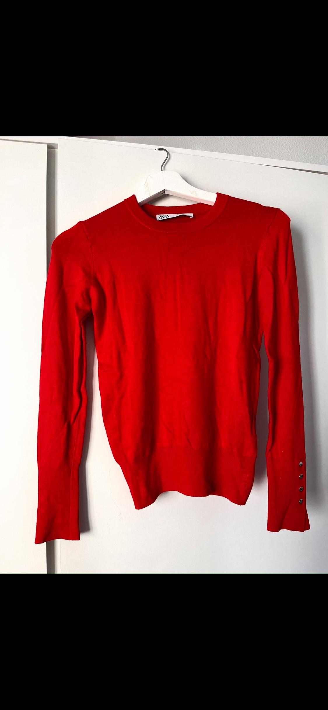 Red long-sleeved jumper hanging on a white hanger, with buttons on the right cuff, against a white background.