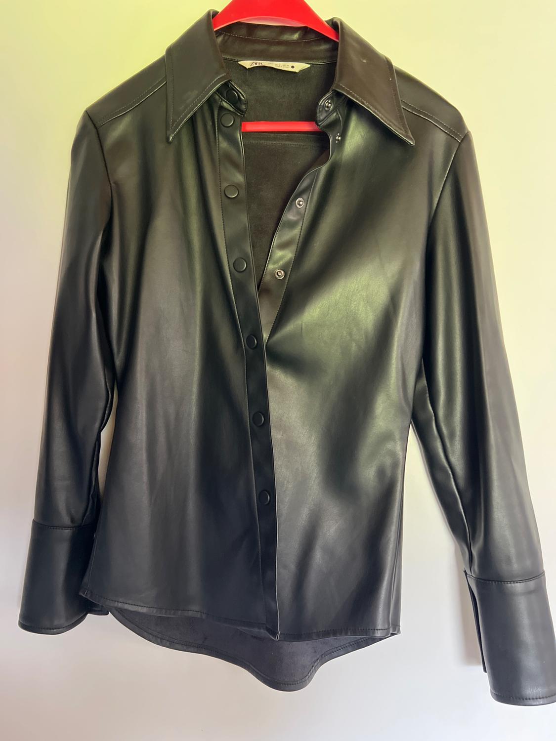 Black faux leather shirt, long-sleeved with press-stud buttons, hanging on a red hanger against a light background. Text: ZARA.