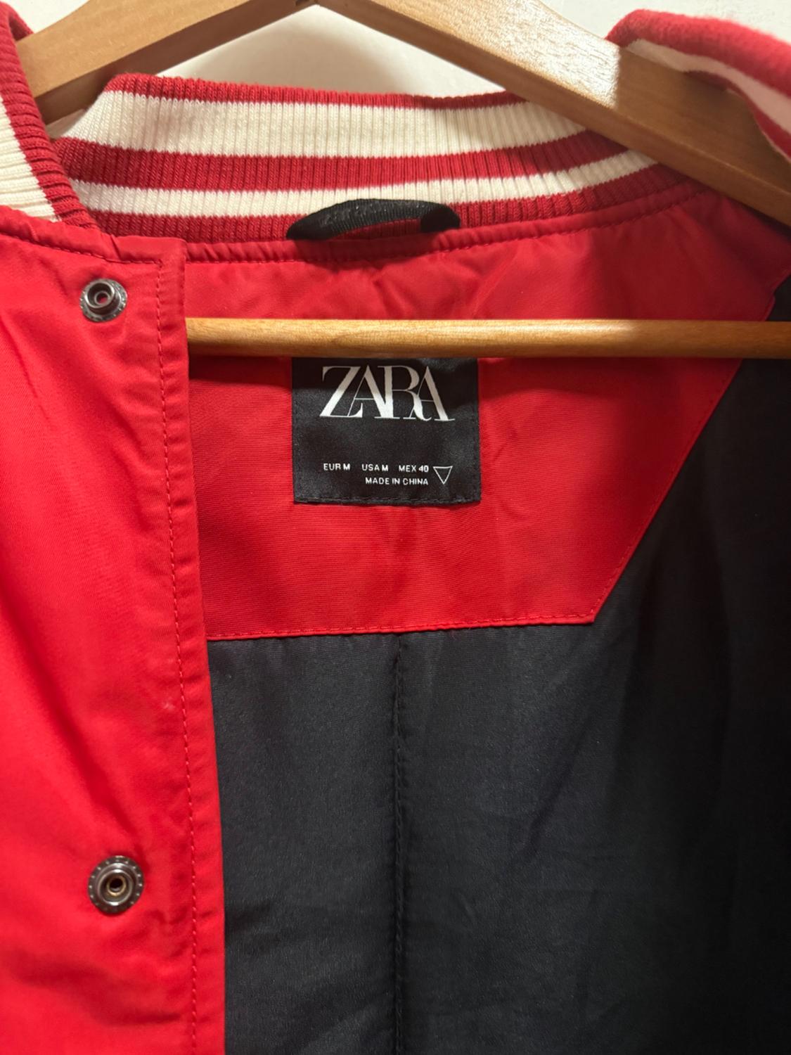 Label of a red jacket with a black interior, white and red striped collar, hanging on a wooden hanger. Visible text: "ZARA".