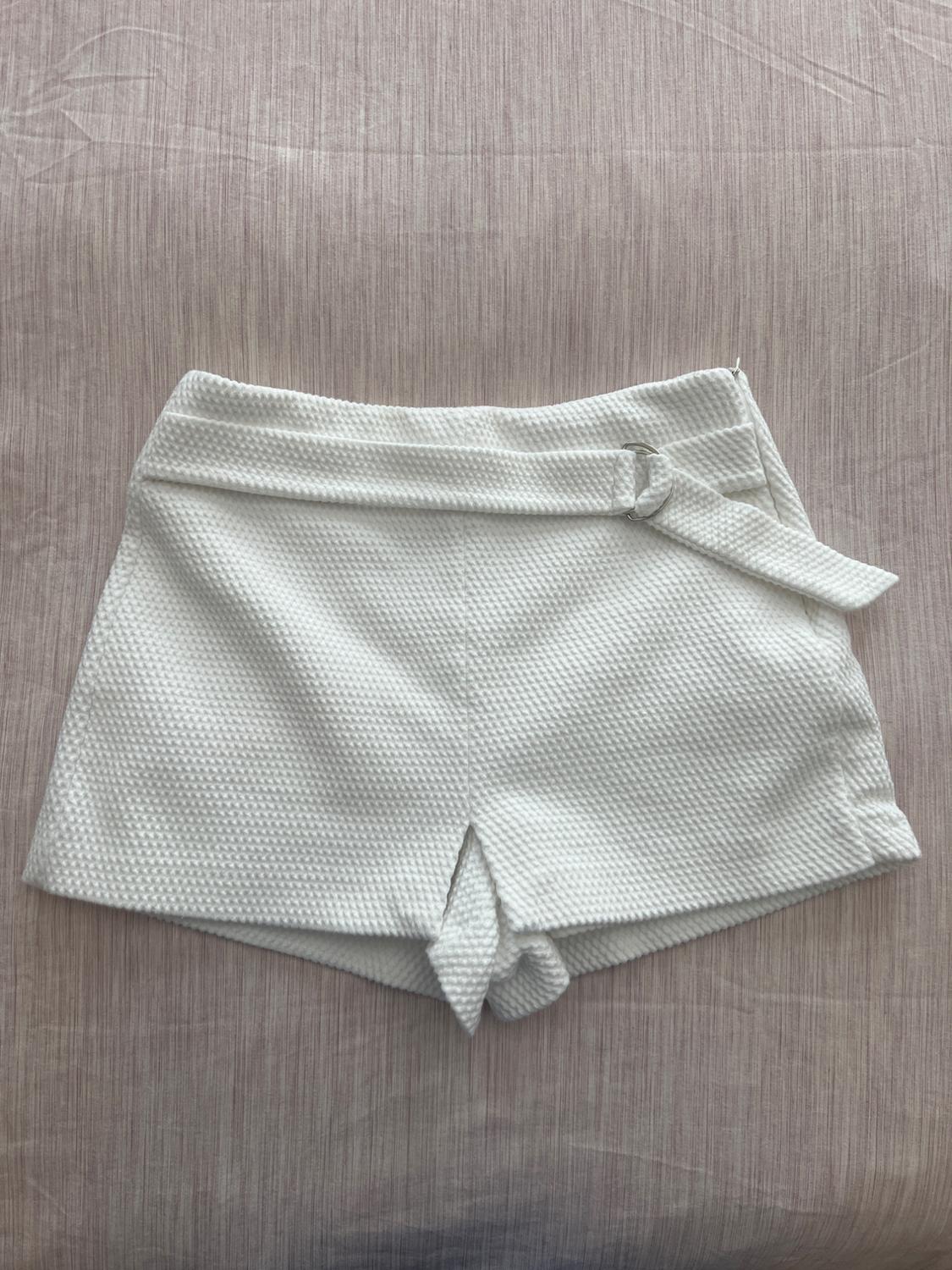 White textured fabric shorts with side belt, on a beige fabric background with subtle lines.