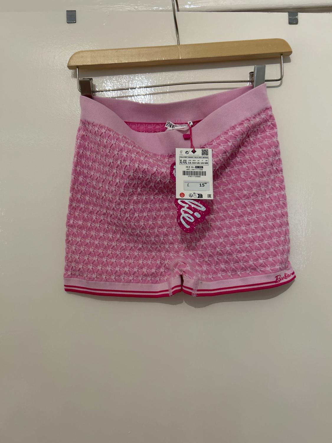 Pink shorts with geometric pattern hanging on a hanger. Tag with price of £15 and text "Barbie" in the bottom right.