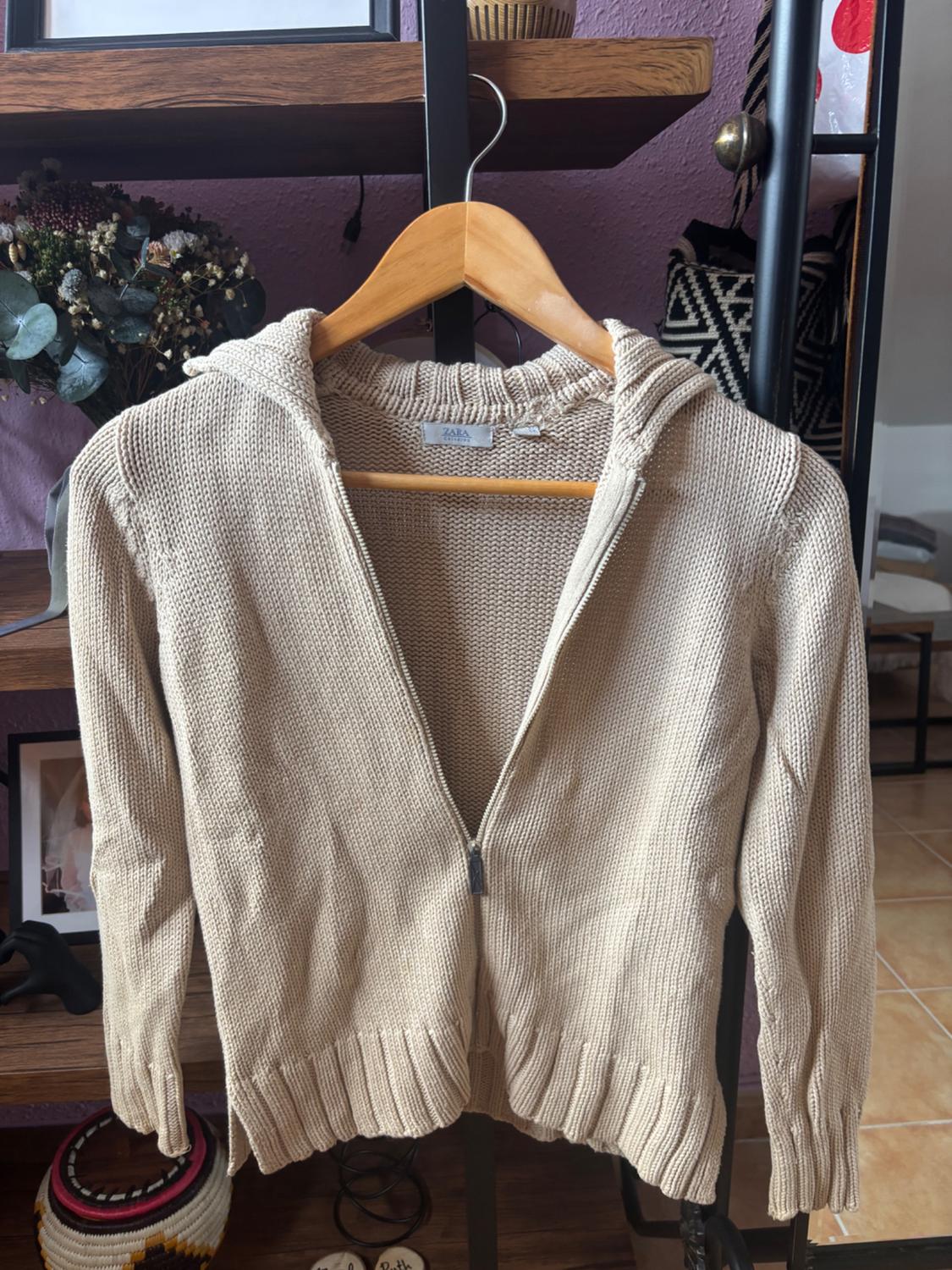 Beige knitted hooded zip-up cardigan hanging on a wooden hanger, with a bookshelf background featuring decorations.