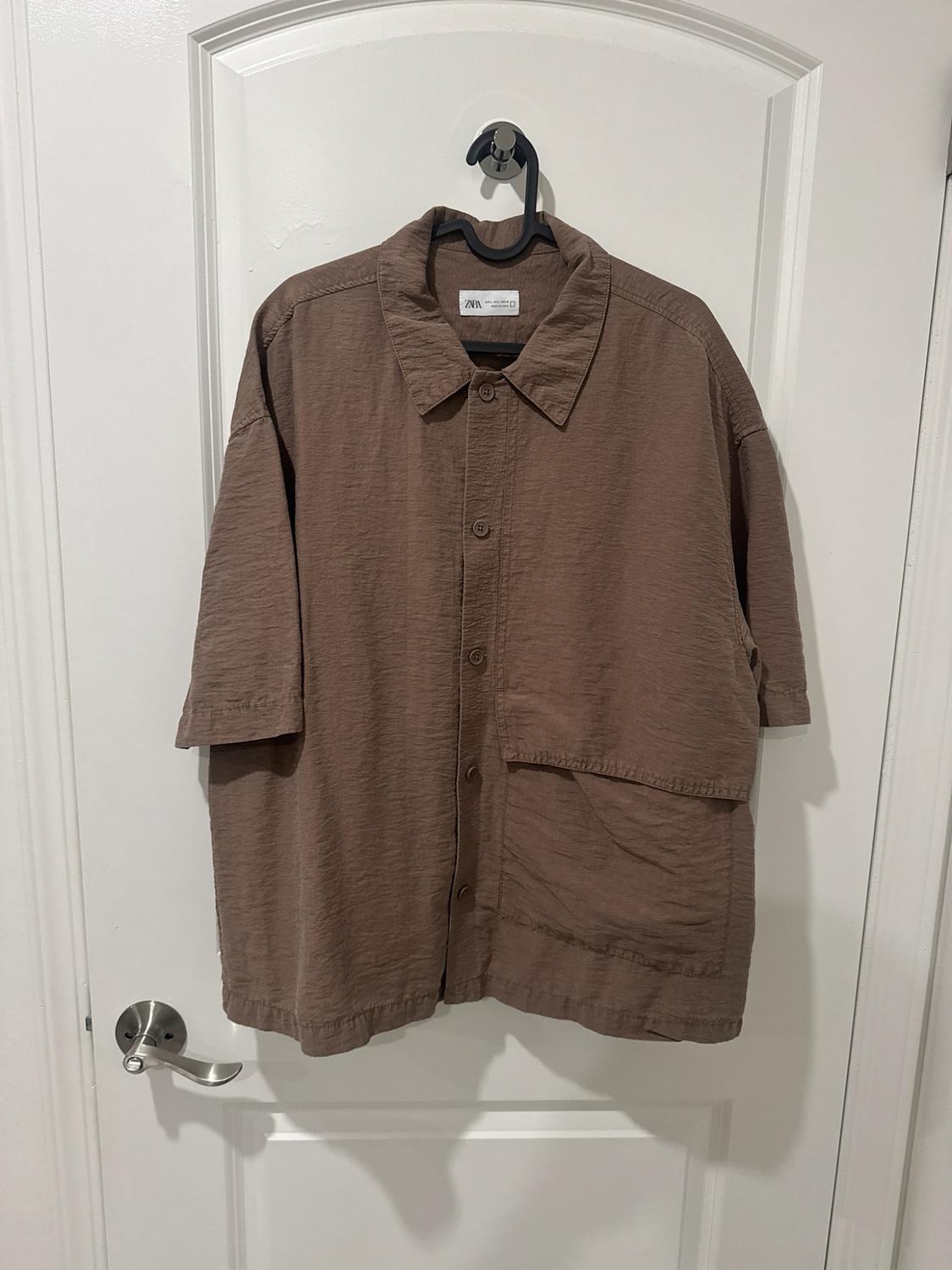 Textured brown short-sleeve shirt with buttons, hanging on a white door. A large pocket and the ZARA label stand out.