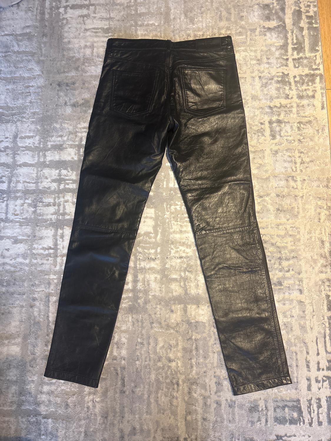 Black leather trousers viewed from behind, placed on a grey rug with a geometric pattern.