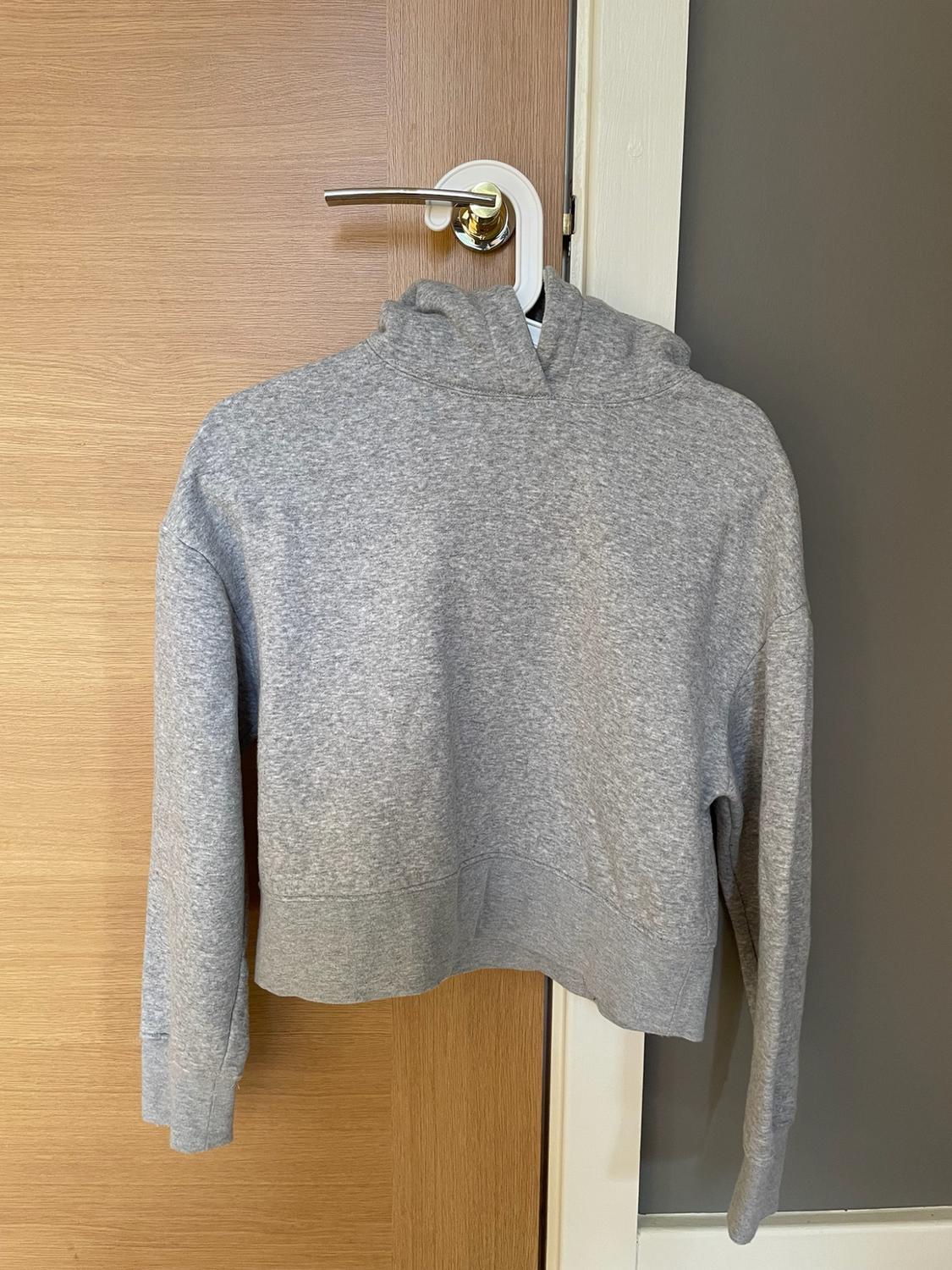 Grey cropped casual hoodie hanging from a hanger on a wooden door handle.
