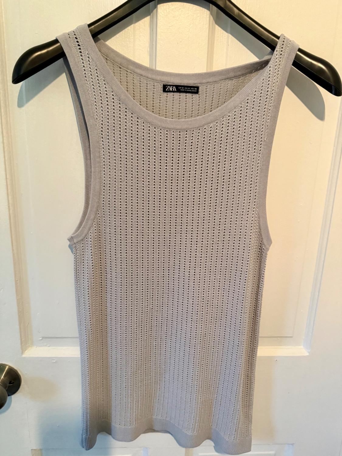 Grey sleeveless top with openwork fabric hanging on a black hanger. Visible label with the text "ZARA". White door in the background.