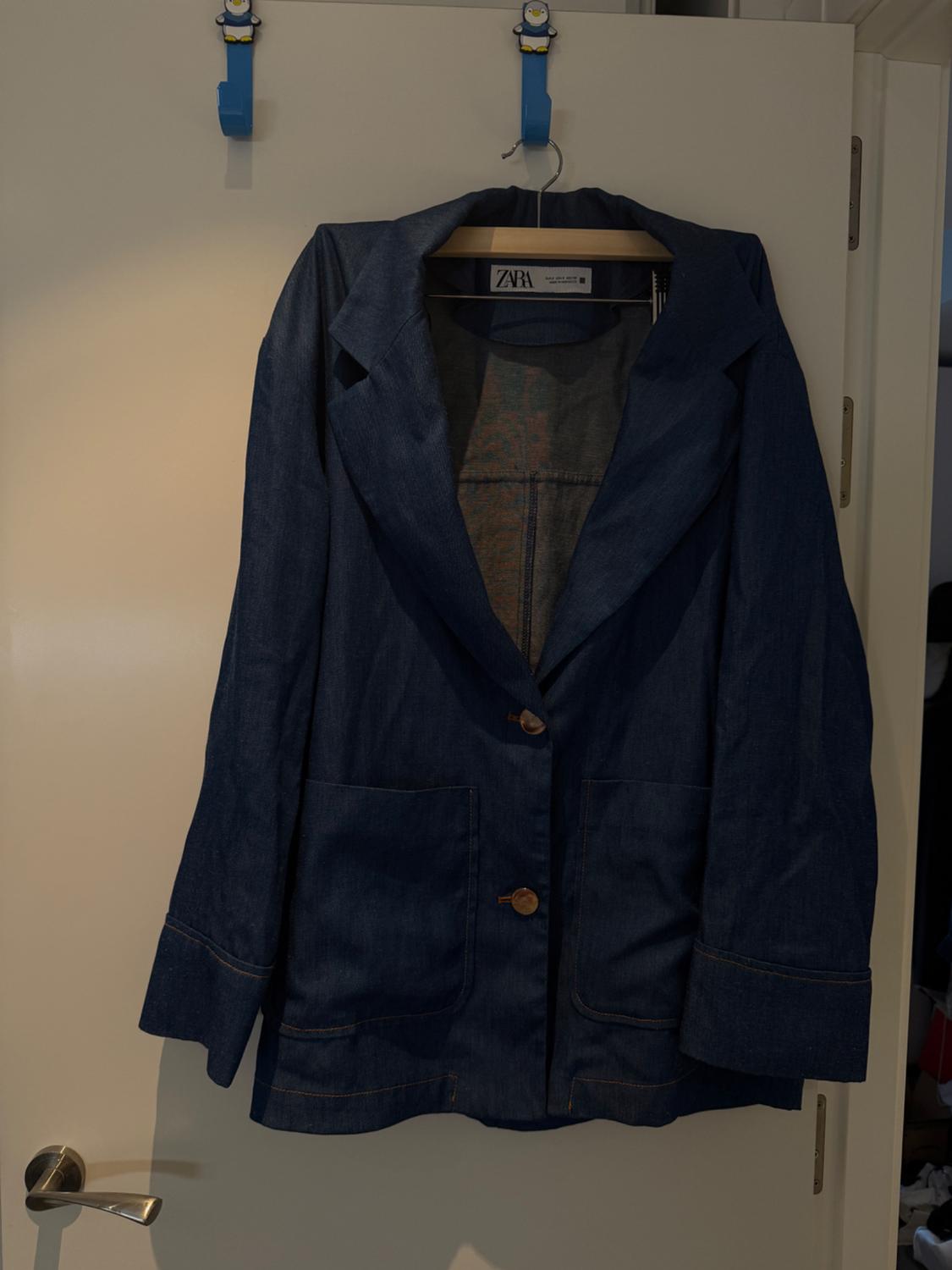 Blue denim jacket hanging on a white door with a wooden hanger. Visible tag: "ZARA". Blue hook with a penguin.